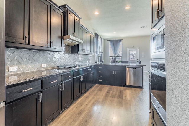 kitchen with decorative backsplash, appliances with stainless steel finishes, dark stone countertops, a peninsula, and under cabinet range hood