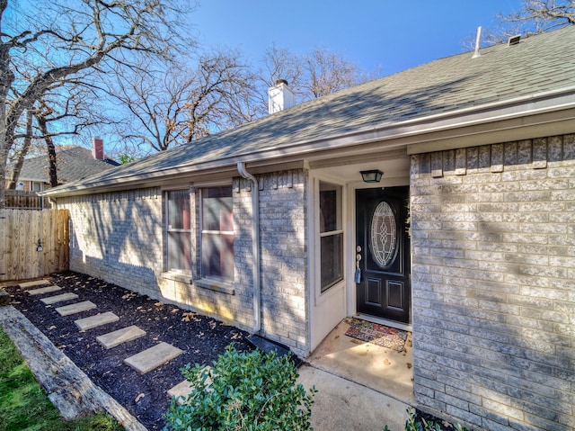 doorway to property featuring roof with shingles, brick siding, a chimney, and fence