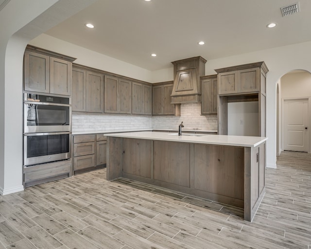 kitchen featuring tasteful backsplash, double oven, sink, custom exhaust hood, and a kitchen island with sink