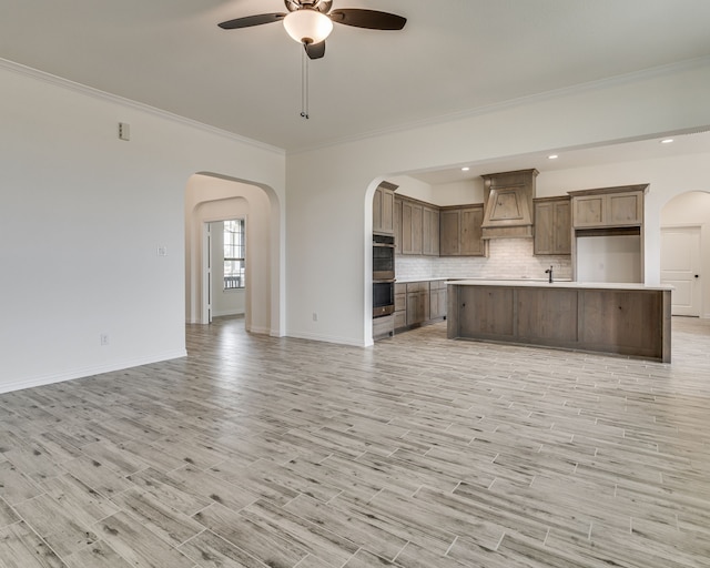 kitchen featuring light hardwood / wood-style flooring, backsplash, custom exhaust hood, stainless steel double oven, and an island with sink