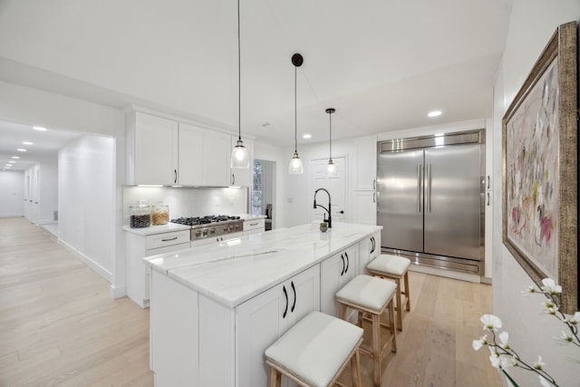 kitchen with a kitchen island with sink, stainless steel appliances, hanging light fixtures, light stone countertops, and white cabinets