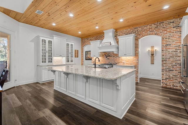 kitchen with arched walkways, premium range hood, white cabinetry, light stone countertops, and a center island with sink