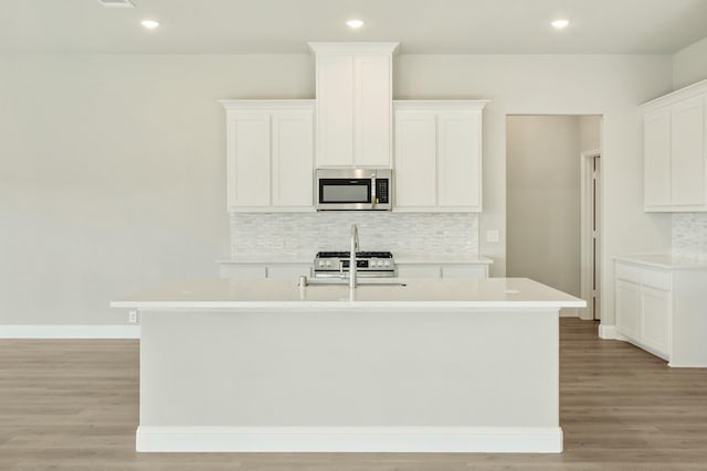 kitchen featuring light countertops, stainless steel microwave, light wood finished floors, a center island with sink, and decorative backsplash