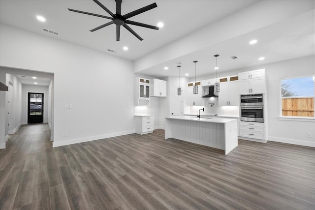 kitchen with a center island with sink, hanging light fixtures, white cabinets, wall chimney range hood, and sink