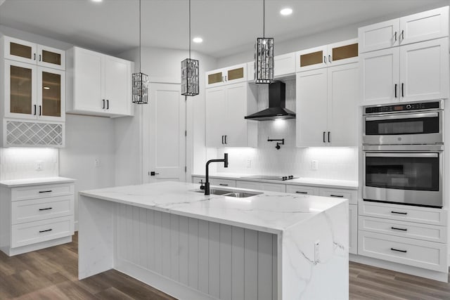 kitchen featuring a center island with sink, light stone counters, wall chimney range hood, sink, and white cabinetry