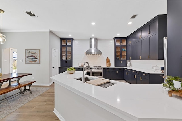 kitchen with visible vents, light countertops, wall chimney range hood, hanging light fixtures, and glass insert cabinets