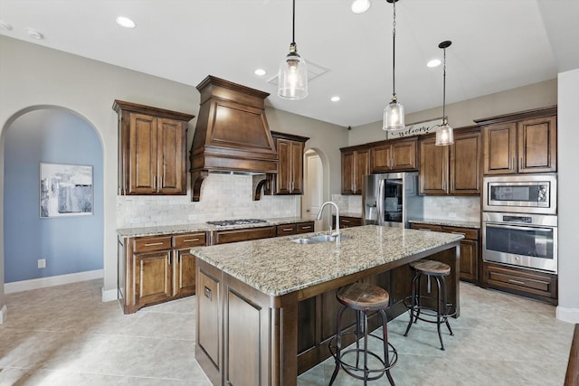 kitchen with arched walkways, light stone counters, stainless steel appliances, premium range hood, and a center island with sink