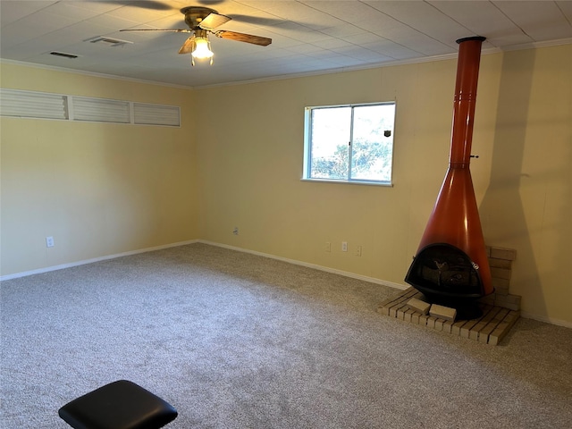 carpeted empty room with a wood stove, ceiling fan, and ornamental molding