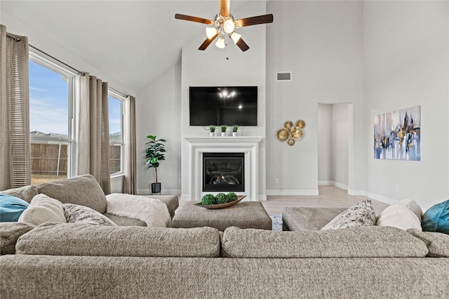 living room with a ceiling fan, visible vents, high vaulted ceiling, baseboards, and a glass covered fireplace