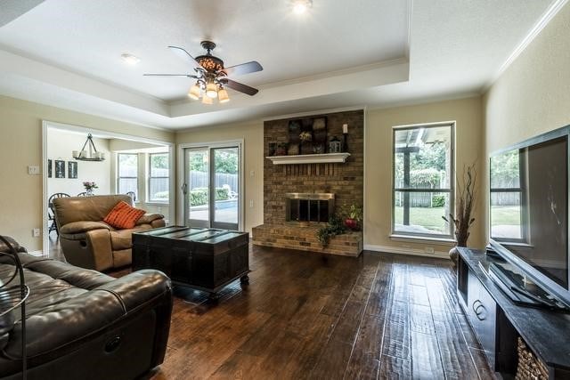 living room featuring a brick fireplace, dark hardwood / wood-style flooring, ceiling fan, a raised ceiling, and crown molding