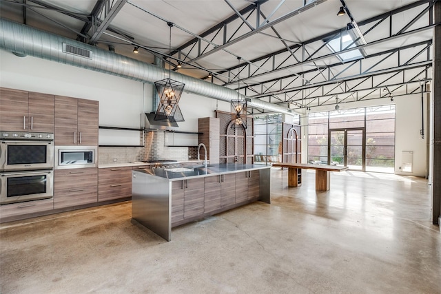 kitchen with a high ceiling, a kitchen island with sink, stainless steel appliances, and hanging light fixtures