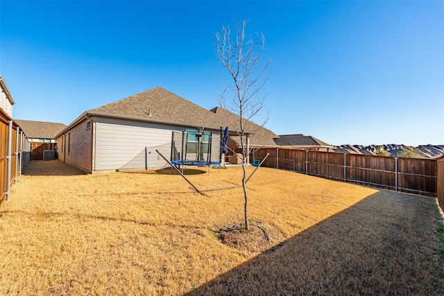 back of property with a trampoline, brick siding, a shingled roof, a lawn, and a fenced backyard