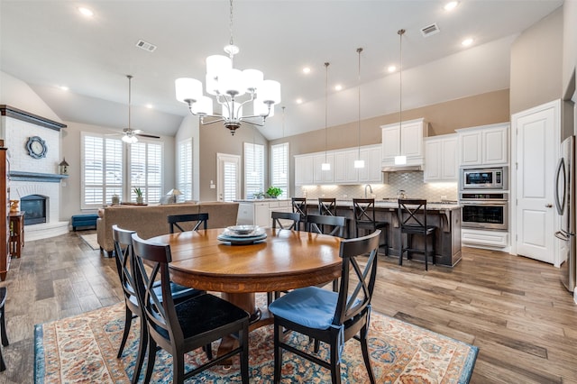 dining area featuring a fireplace, high vaulted ceiling, wood-type flooring, and ceiling fan with notable chandelier