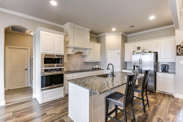 kitchen with an island with sink, white cabinetry, stainless steel appliances, sink, and dark stone counters