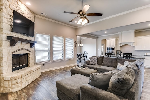 living room featuring ceiling fan with notable chandelier, ornamental molding, a stone fireplace, and wood-type flooring