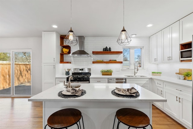 kitchen with stainless steel appliances, a sink, wall chimney exhaust hood, open shelves, and an island with sink