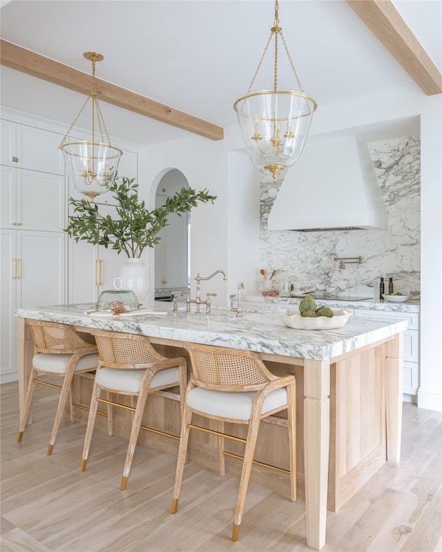 kitchen featuring an island with sink, tasteful backsplash, custom range hood, beamed ceiling, and a chandelier