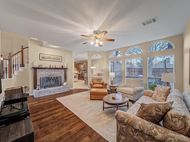 living room featuring a textured ceiling, wood finished floors, visible vents, a ceiling fan, and a brick fireplace