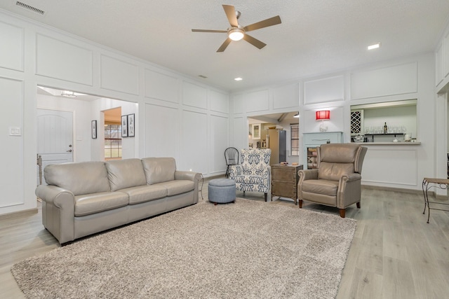 living room featuring ceiling fan, a decorative wall, visible vents, light wood-style floors, and crown molding