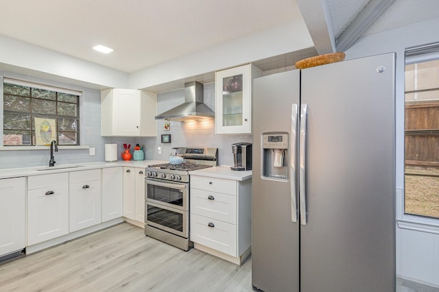 kitchen with a sink, light countertops, appliances with stainless steel finishes, wall chimney exhaust hood, and glass insert cabinets