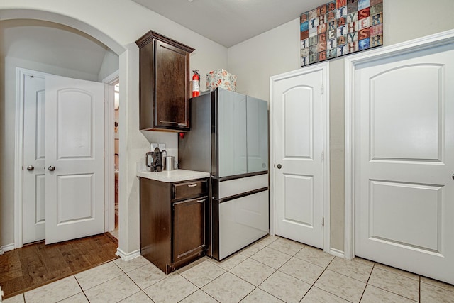 kitchen featuring dark brown cabinetry, light countertops, freestanding refrigerator, light tile patterned flooring, and arched walkways