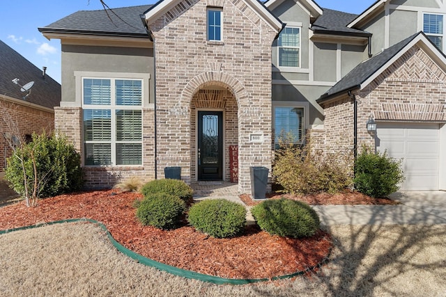 view of front of house featuring brick siding, roof with shingles, an attached garage, and stucco siding