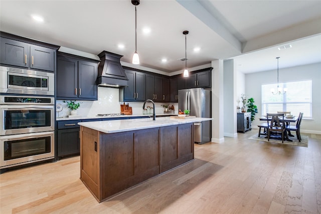 kitchen with premium range hood, pendant lighting, stainless steel appliances, and a kitchen island with sink