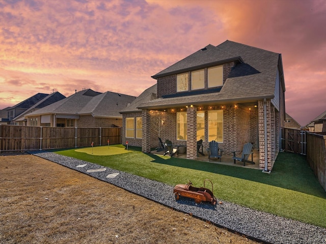 back of house at dusk featuring a shingled roof, brick siding, a patio, and a fenced backyard