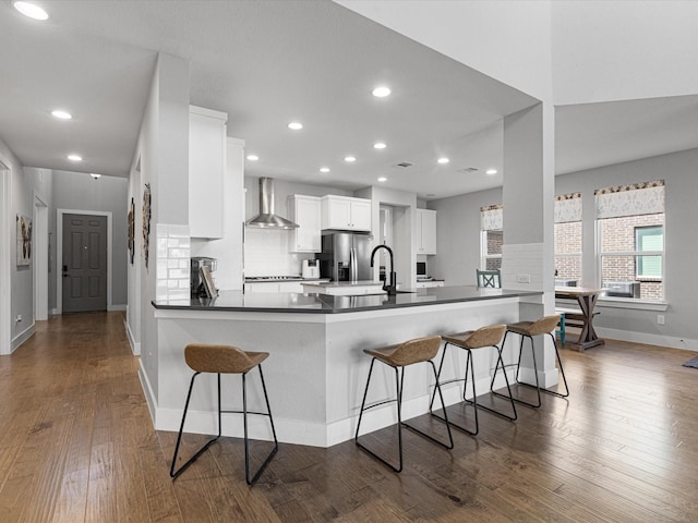 kitchen with a peninsula, white cabinetry, a kitchen breakfast bar, wall chimney exhaust hood, and stainless steel fridge