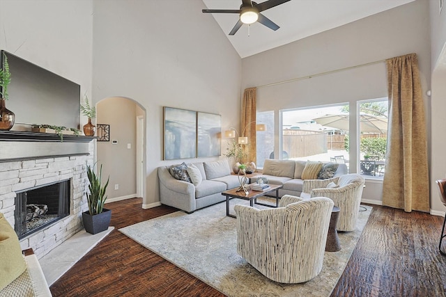 living area featuring arched walkways, ceiling fan, a stone fireplace, wood finished floors, and high vaulted ceiling