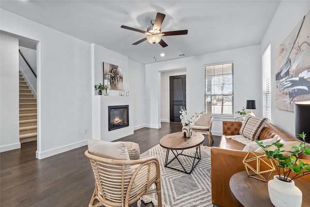 living room featuring dark wood-style floors, a glass covered fireplace, ceiling fan, baseboards, and stairs