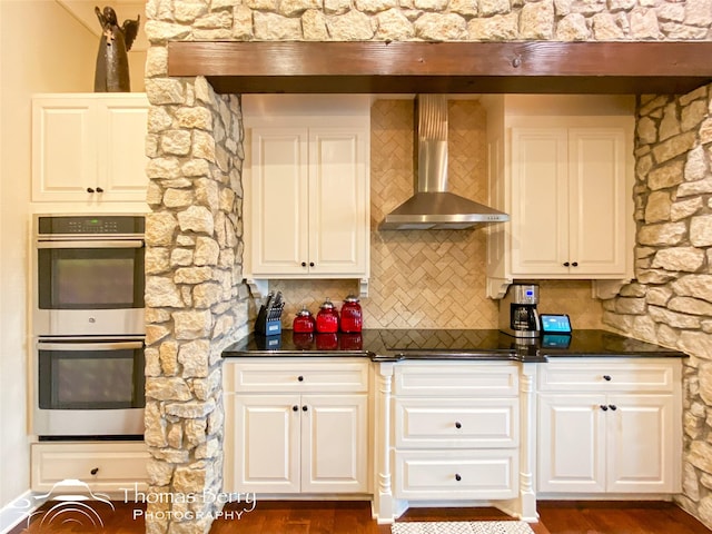 kitchen featuring double oven, wall chimney range hood, and white cabinets