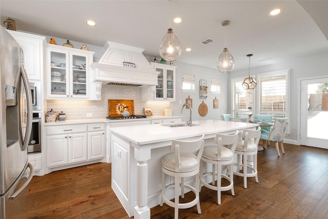 kitchen featuring premium range hood, white cabinets, glass insert cabinets, light countertops, and a center island with sink