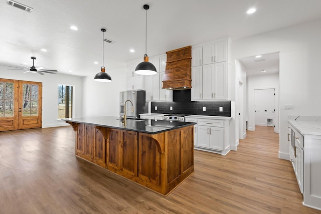 kitchen featuring hanging light fixtures, visible vents, a center island with sink, white cabinets, and custom exhaust hood