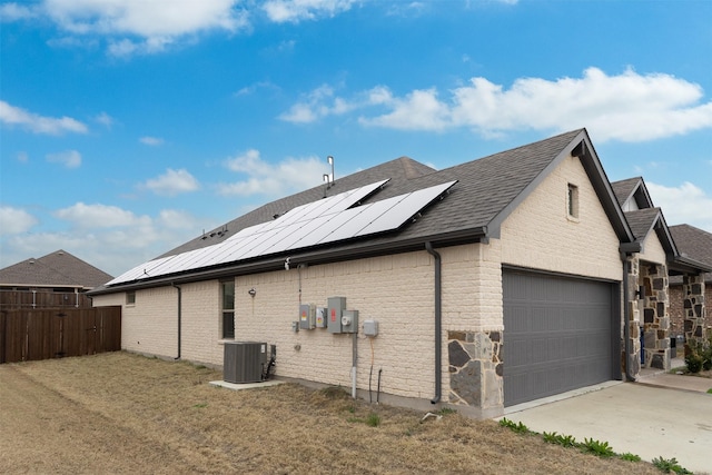 view of side of home with brick siding, a shingled roof, central air condition unit, roof mounted solar panels, and fence