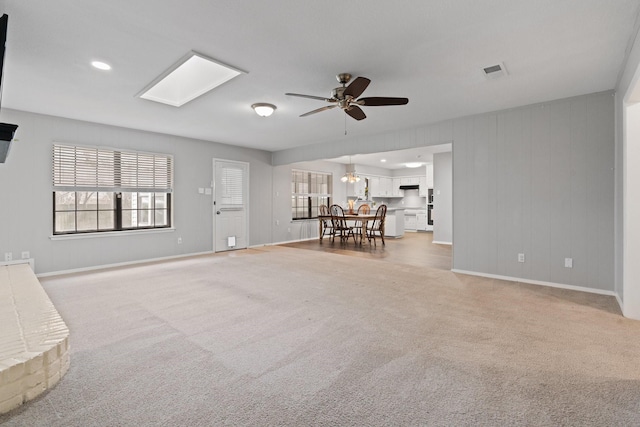 unfurnished living room with a skylight, visible vents, a ceiling fan, baseboards, and light carpet