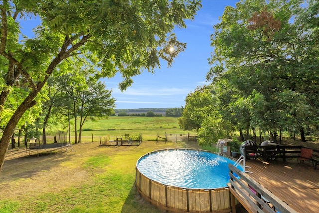 view of swimming pool featuring a lawn, a trampoline, fence, a rural view, and a wooden deck