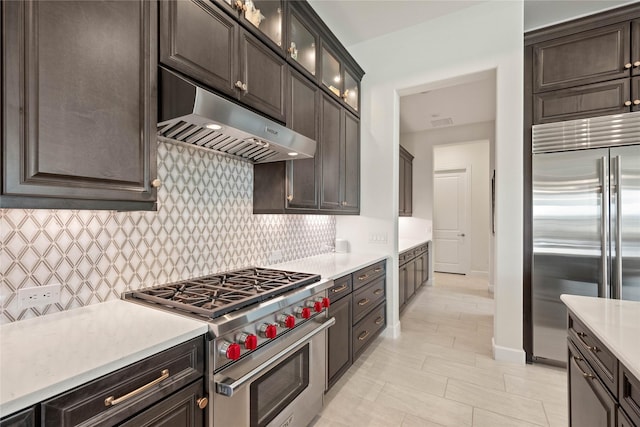 kitchen featuring tasteful backsplash, premium appliances, glass insert cabinets, light countertops, and under cabinet range hood