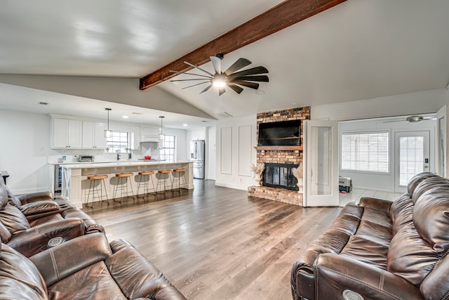 living area with ceiling fan, lofted ceiling with beams, baseboards, light wood-type flooring, and a brick fireplace