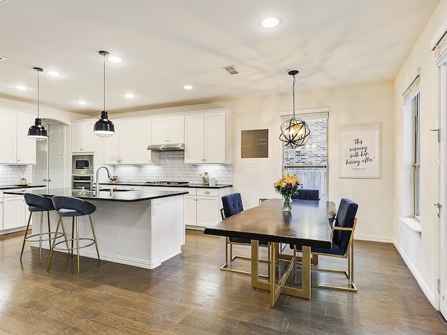 kitchen with dark countertops, a center island with sink, white cabinets, and hanging light fixtures