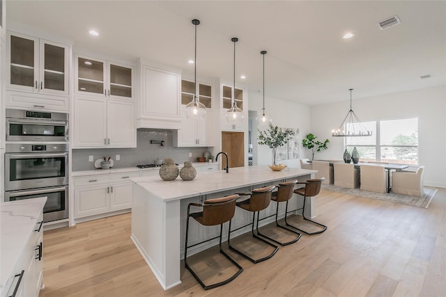 kitchen featuring light wood-style flooring, a kitchen island with sink, light stone countertops, visible vents, and decorative backsplash