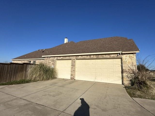 view of side of property with brick siding, roof with shingles, an attached garage, fence, and driveway