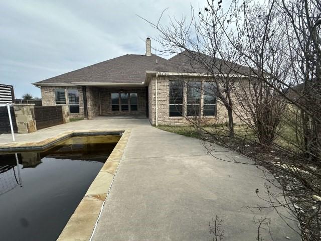 back of house with brick siding, a shingled roof, driveway, a chimney, and a patio area