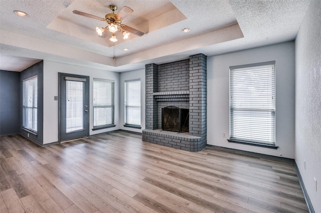 unfurnished living room featuring a raised ceiling, a ceiling fan, wood finished floors, a textured ceiling, and a brick fireplace
