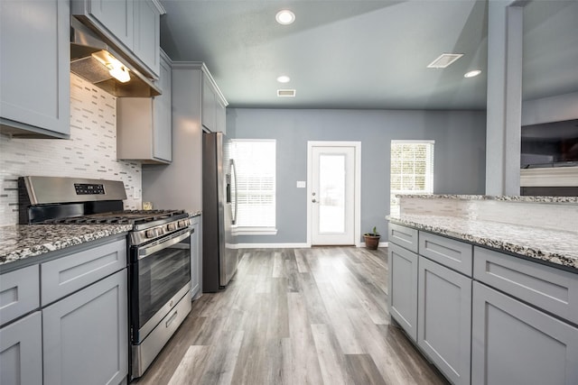 kitchen featuring light stone counters, under cabinet range hood, gray cabinetry, appliances with stainless steel finishes, and backsplash