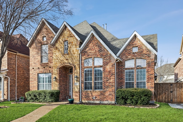 tudor home featuring brick siding, roof with shingles, fence, stone siding, and a front lawn