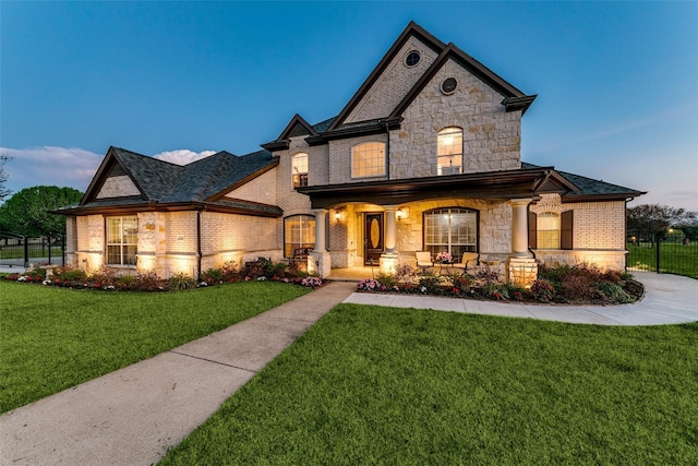 french country style house with covered porch, brick siding, fence, stone siding, and a front lawn