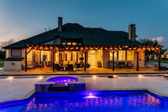 back of house at dusk with brick siding, a pool with connected hot tub, an outdoor living space, a chimney, and a patio area