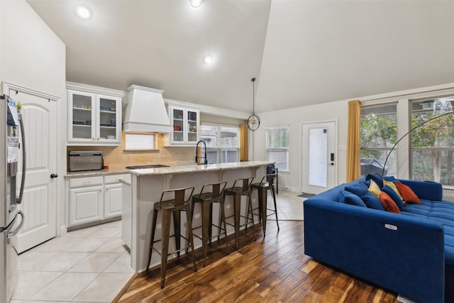 kitchen with white cabinets, an island with sink, lofted ceiling, custom range hood, and glass insert cabinets