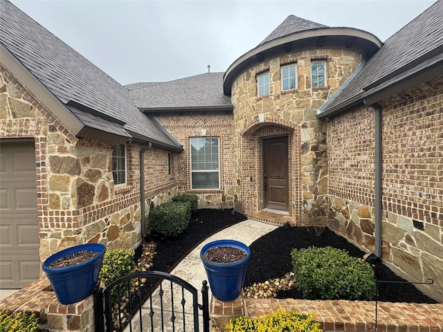 view of exterior entry with stone siding, roof with shingles, an attached garage, a gate, and brick siding